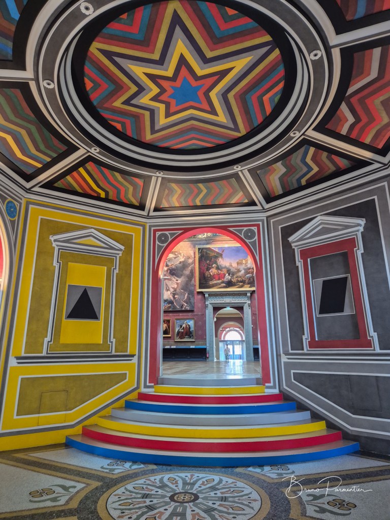 Vue intérieure d'un espace coloré avec un plafond étoilé, des murs décorés et un escalier multicolore menant à un couloir avec des tableaux.