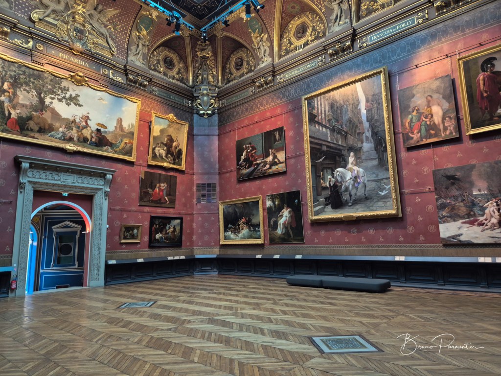 Intérieur d'un musée avec des peintures accrochées aux murs, le sol en bois en chevrons et un éclairage artistiquement orienté.