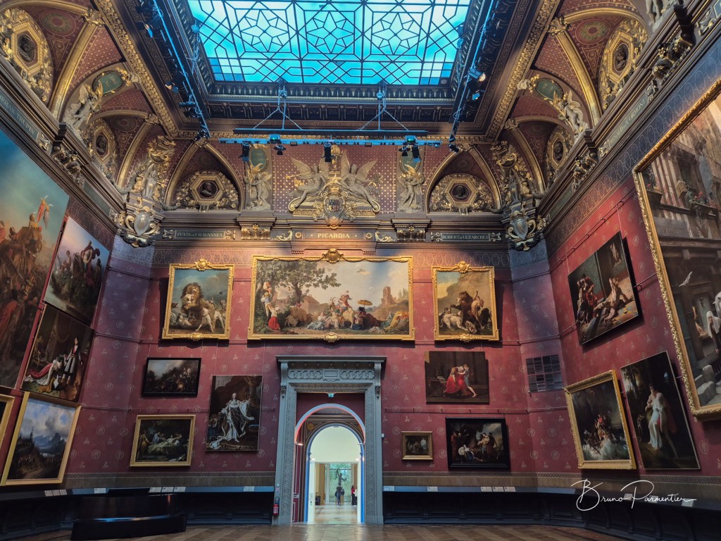 Intérieur d'une galerie d'art avec des œuvres célèbres accrochées aux murs et un plafond orné de détails architecturaux.